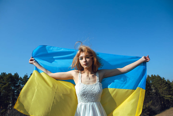 Πορτρέτο της γυναίκας τυλιγμένο σε ουκρανική κίτρινη μπλε σημαία κυματίζει στον άνεμο. Εθνικό σύμβολο της Ουκρανίας. Πατριωτική κυρία με μακιγιάζ που περπατάει στο χωράφι. Σταθείτε με την Ουκρανία, υποστήριξη και αλληλεγγύη - Φωτογραφία, εικόνα