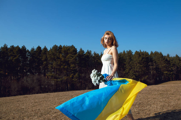 Portrait of woman wrapped in ukrainian yellow blue flag flutters in the wind. National symbol of Ukraine. Patriotic lady with makeup and chamomile flowers. Stand with Ukraine, support and solidarity - Foto, imagen