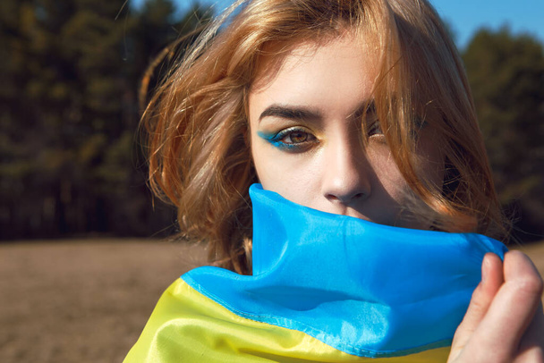 Portrait of woman wrapped in ukrainian yellow blue flag flutters waving in the wind. National symbol of Ukraine. Patriotic lady with makeup walking in the field. Stand with Ukraine, support and solidarity - Foto, imagen