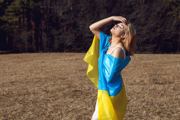 Portrait of woman wrapped in ukrainian yellow blue flag flutters waving in the wind. National symbol of Ukraine. Patriotic lady with makeup walking in the field. Stand with Ukraine, support and solidarity - Foto, imagen