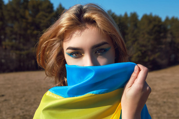 Portrait of woman wrapped in ukrainian yellow blue flag flutters waving in the wind. National symbol of Ukraine. Patriotic lady with makeup walking in the field. Stand with Ukraine, support and solidarity - Φωτογραφία, εικόνα