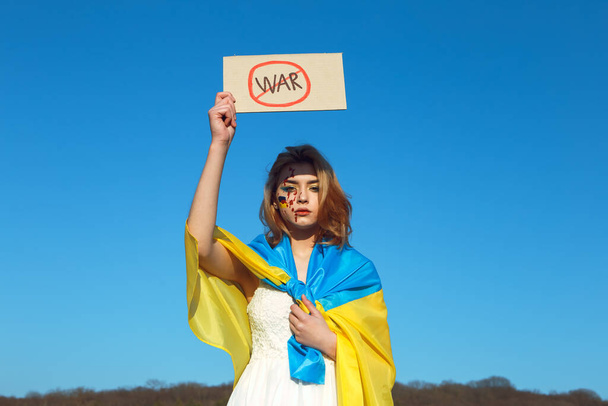 Nessuna guerra! Donna avvolta nella bandiera blu gialla ucraina con cartello di protesta. Simbolo nazionale dell'Ucraina. La signora con il trucco di sangue ha una targa patriottica. Sostegno all'Ucraina, sostegno e solidarietà.  - Foto, immagini