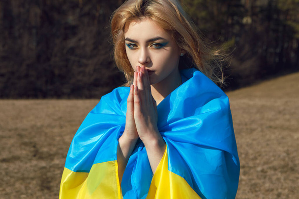 Portrait of woman wrapped in ukrainian yellow blue flag. National symbol of Ukraine. Patriotic lady with makeup show praying gesture sign. Stand with Ukraine, support and solidarity - Photo, Image