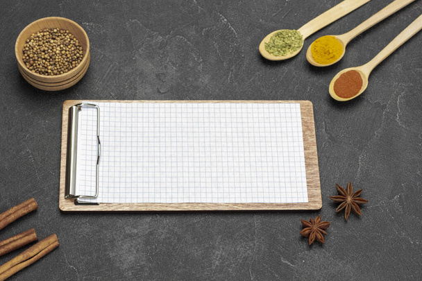 Board-tablet with blank sheet. Three spoons with spices. Cinnamon sticks and coriander seeds in bowl. Flat lay. Black background - Фото, зображення