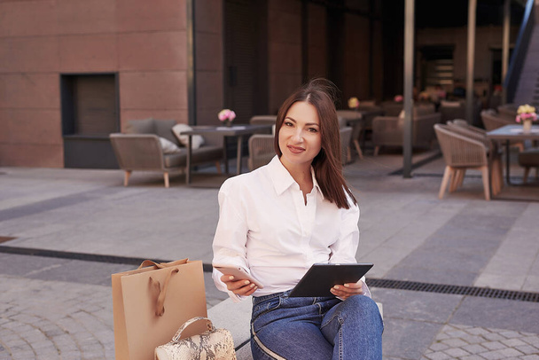 Young beautiful woman walks in city. Portrait of girl on street with phone and tablet. Purchase packages and shopping. Young beautiful woman walks in city. Online work, distance learning - Photo, image