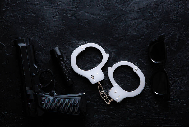 Black gun, flashlight, glasses and Police metal real handcuffs lie on the black background. Private detective work. Searching information. Crime and robbery, prison concept. - Photo, Image