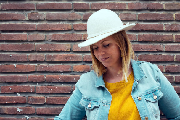 caucasian blond woman wearing white hat - denim jacket - yellow sweater against a brick wall background - Foto, Imagen