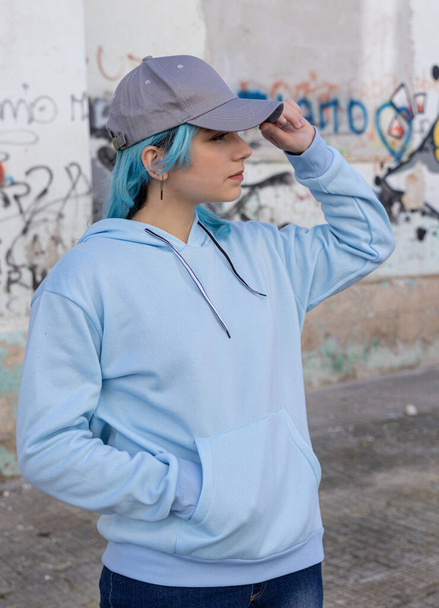 Γαλαζομάλλης έφηβη με μπλε κουκούλα και καπέλο του μπέιζμπολ να αγγίζει το καπέλο της και να κοιτάζει δεξιά. Μπλε μαλλιά έφηβος κορίτσι μένει σε εξωτερικούς χώρους κατά γκράφιτι τοίχο. Ενδύματα και καπάκια  - Φωτογραφία, εικόνα