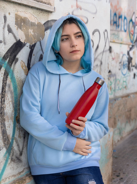 Έφηβος με μπλε κουκούλα και κόκκινο μπουκάλι με θέα στην άκρη. Μπλε μαλλιά έφηβος κορίτσι που διαμένουν έξω κοντά στον τοίχο γκράφιτι. Ρούχα και επαναχρησιμοποιήσιμα καλλυντικά μπουκαλιών. Έννοια hipster και εφηβείας - Φωτογραφία, εικόνα