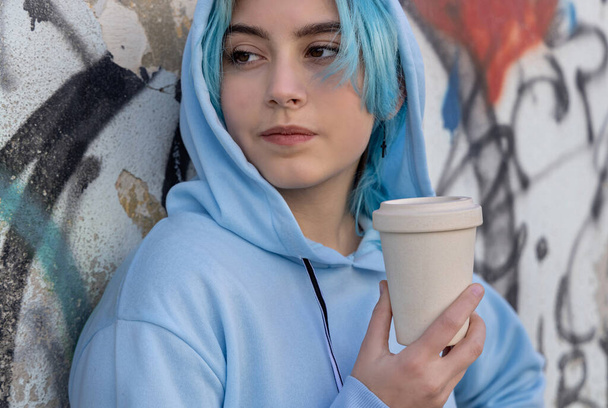 Εφηβικό κορίτσι σε γαλάζιο, υπερμεγέθη φούτερ με καφέ για να πάει και να κοιτάξει στην άκρη. Μπλε μαλλιά έφηβος κορίτσι που διαμένουν έξω κοντά στον τοίχο γκράφιτι. Απομίμηση κυπέλλου. Έννοια hipster και εφηβείας - Φωτογραφία, εικόνα