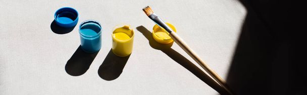 high angle view of blue and yellow paint in jars near paintbrush on white, banner - Photo, Image