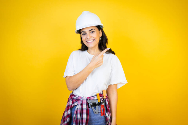Nuori valkoihoinen nainen yllään kypärä ja rakentaja vaatteita yli eristetty keltainen tausta hymyilee ja osoittaa käsin ja sormella sivulle - Valokuva, kuva