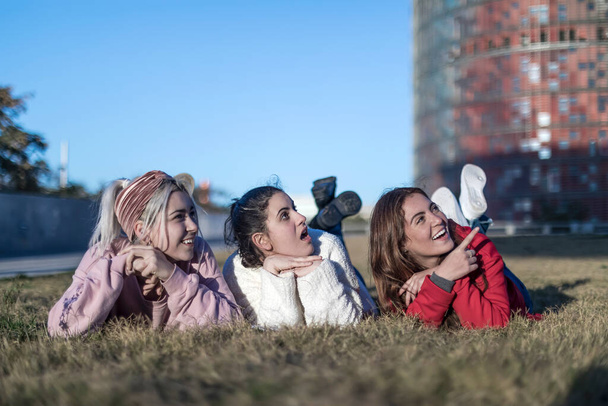 Three happy female friends lying on the grass in a public park at the city - Foto, imagen