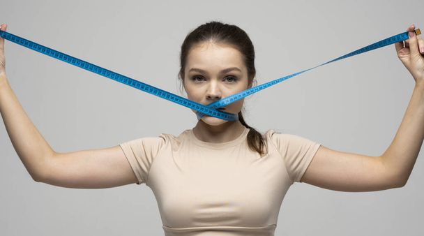 Young attractive beautiful brunette woman in a beige outfit holding measuring tape around her mouth on white background. Healthy lifestyle and diet concept. - Fotó, kép
