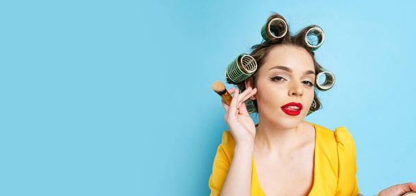 Comic portrait of young emotional girl, housewife with curlers on her head isolated on blue studio background. Emotions, female rights, beauty, family - Foto, afbeelding