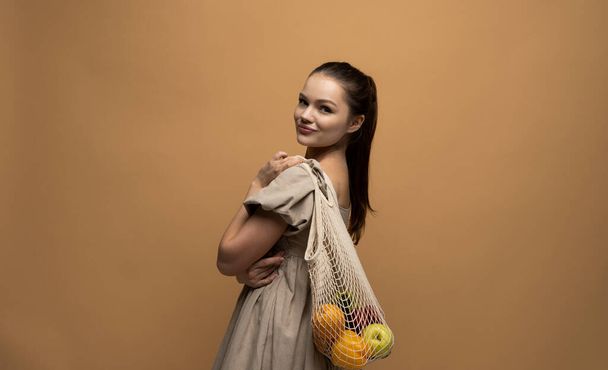 Side view of attractive happy brunette woman holding mesh grocery bag with vegetables. Concept of no plastic. Zero waste, plastic free. Eco friendly concept. Sustainable lifestyle. - Photo, image