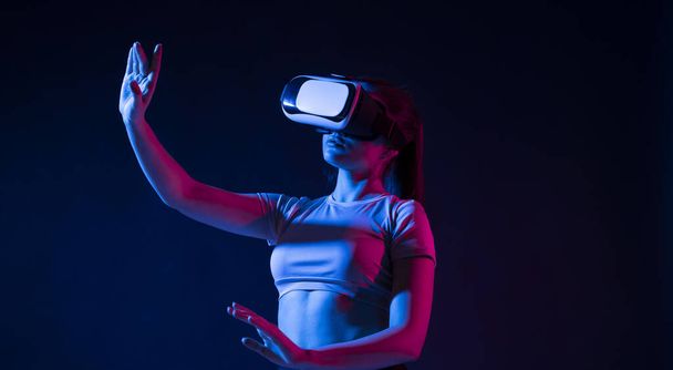 Modern architect using virtual reality glasses at workplace and touch something in virtual world. Woman working in VR goggles in neon light. Designer working in VR studio. - 写真・画像