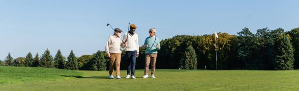 senior interracial friends walking with golf clubs on green field, banner - 写真・画像