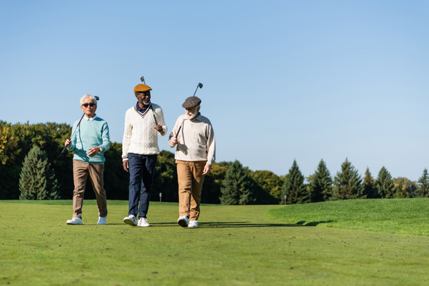 senior interracial amigos caminando con golf clubs en verde campo - Foto, Imagen