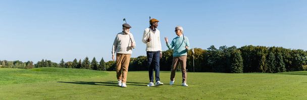 senior interracial friends walking with golf clubs on field, banner - Foto, Imagem