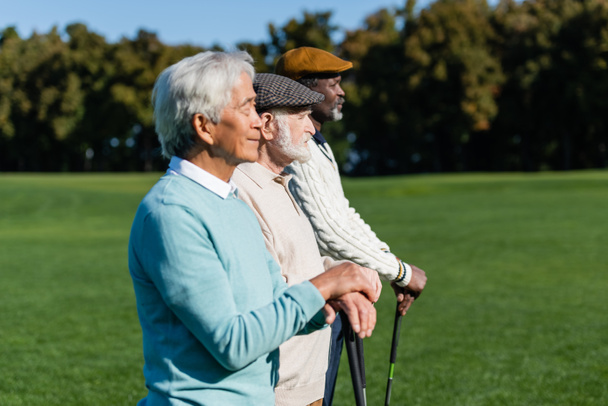Seitenansicht eines älteren Mannes mit Schiebermütze in der Nähe von Freunden mit Golfschlägern - Foto, Bild
