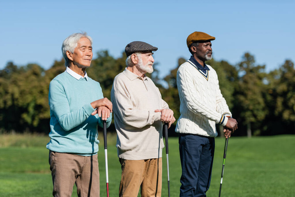 senior man in flat cap near interracial friends standing with golf clubs - Photo, Image
