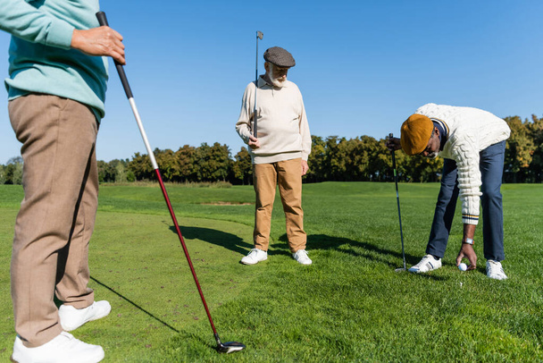 Afrikanisch-amerikanischer Senior platziert Ball auf Golfabschlag in der Nähe multiethnischer Freunde  - Foto, Bild