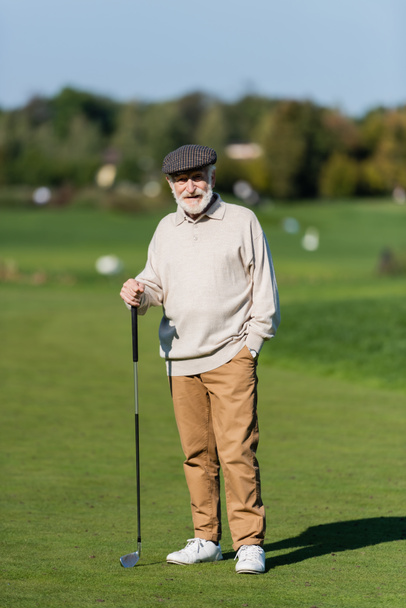 uomo anziano con berretto piatto in piedi con mano in tasca e golf club su campo verde  - Foto, immagini