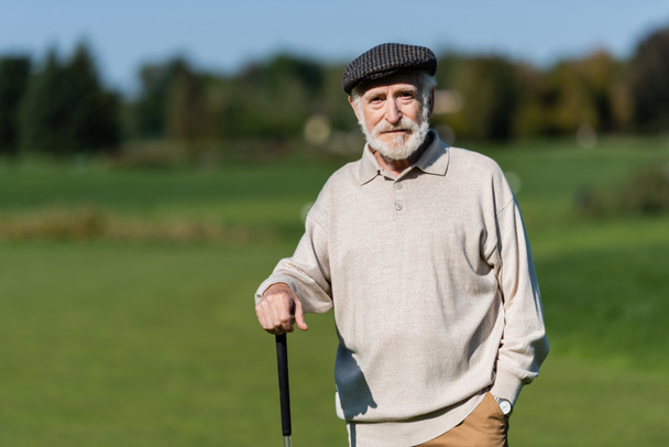 bohatý starší muž v ploché čepici stojící s golfovou holí - Fotografie, Obrázek