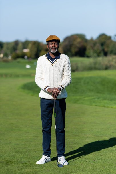 Orta yaşlı Afro-Amerikan bir adam elinde golf sopasıyla yeşil alanda duruyor. - Fotoğraf, Görsel