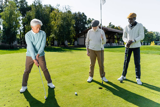senior man in flat cap holding flag stick near hole and multiethnic friends  - Photo, Image