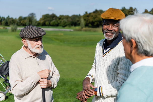 seniores multiétnicos homens segurando tacos de golfe e falando com desfocado asiático amigo  - Foto, Imagem