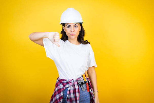 Jonge blanke vrouw draagt hardhoed en bouwer kleding over geïsoleerde gele achtergrond met boos gezicht, negatief teken toont afkeer met duim naar beneden - Foto, afbeelding