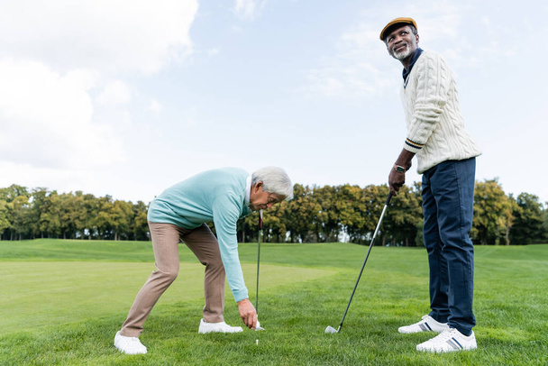 asian senior man putting ball on golf tee near african american friend  - 写真・画像
