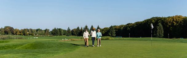 azjatycki mężczyzna w okularach przeciwsłonecznych chodzenie w pobliżu starszych wielonarodowych przyjaciół z kijami golfowymi, sztandar - Zdjęcie, obraz