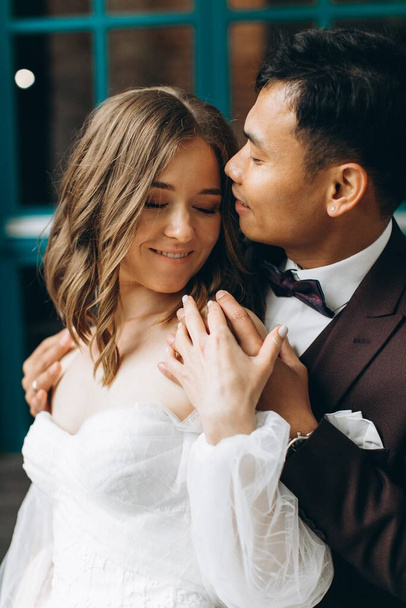 Wedding day. Beautiful European bride and her Asian groom posing against the backdrop of cafes and large windows. - Foto, afbeelding