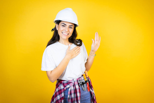 Jonge blanke vrouw draagt hardhoed en bouwer kleding over geïsoleerde gele achtergrond glimlachend met de hand op de borst en vingers omhoog, het maken van een trouw eed - Foto, afbeelding