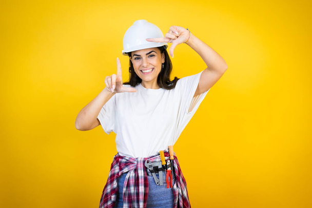 Mujer joven caucásica con sombrero de fuerza y ropa de constructor sobre fondo amarillo aislado sonriendo haciendo marco con manos y dedos con cara feliz - Foto, Imagen