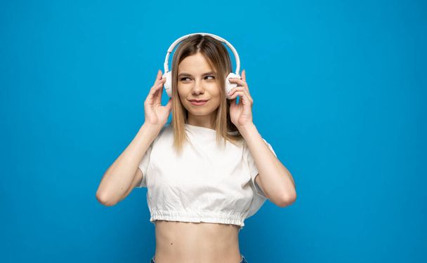 Beautiful young woman in headphones listening to music blue background. Young beautiful woman in bright outfit enjoying a music. - Foto, immagini