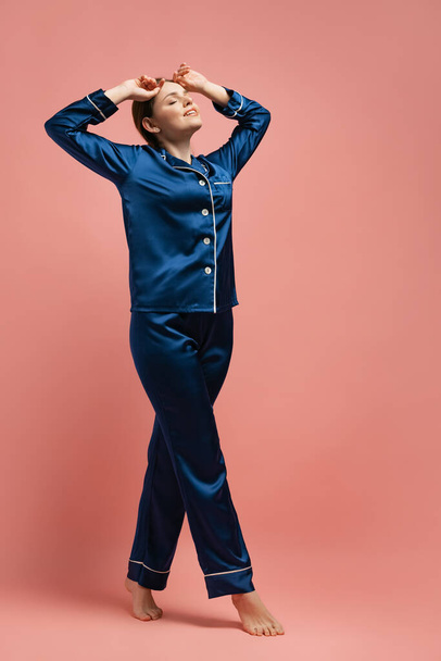 Portrait of young woman in blue silk pajamas posing isolated over pink studio background - Foto, immagini