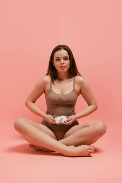 Full-length portrait of young woman in cotton bodysuit sitting with orchid flower on belly, posing isolated over pink studio background - Foto, Imagem