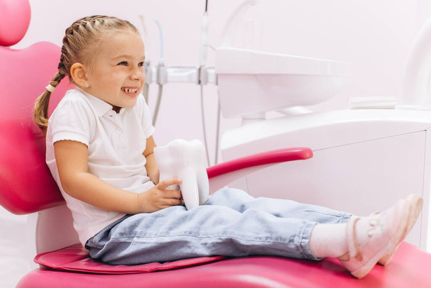 Маленька дівчинка посміхається під час візиту до стоматолога, який тримає модель зуба в стоматологічній клініці
 - Фото, зображення