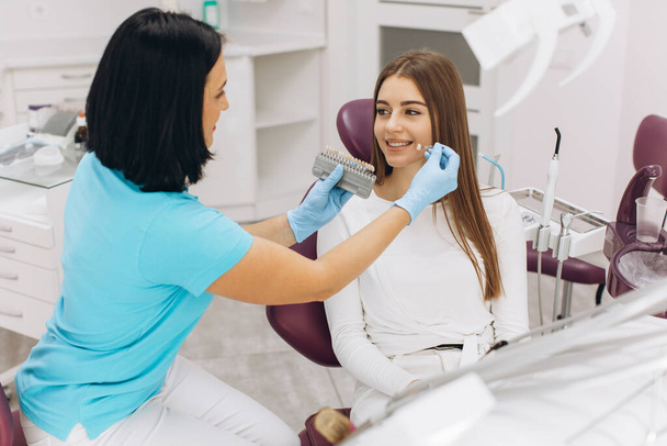 Une dentiste aide sa patiente à choisir la couleur de l'implant ou de la couronne à l'aide d'une échelle de couleurs - Photo, image