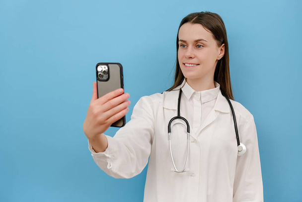 Smiling young female doctor holding phone talk to patient make telemedicine online conference video call, isolated on blue wall. Gp remote consultation telemedicine virtual mobile health care chat app - 写真・画像