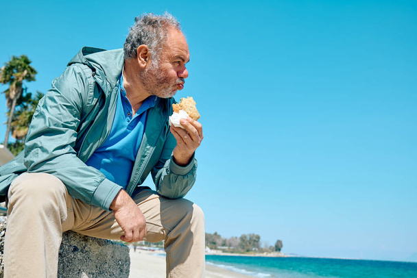 Bearded mature man at spring seaside eating hot palatable panzarotto (calzone pizza fried in hot oil). Typical Sicilian street food. - Fotoğraf, Görsel