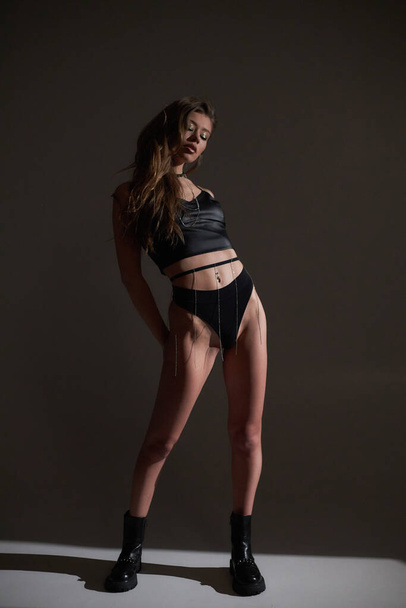 Studio πορτρέτο του νεαρού ελκυστική γυναίκα μοντέλο με μελαχρινή μακριά σγουρά μαλλιά, σε μαύρο φόρεμα bodysuit και ψηλά τακούνια, μακριά πόδια με μαυρισμένο δέρμα, λεπτό σώμα, μείνετε στο παρασκήνιο με ήλιο σκληρό φως. - Φωτογραφία, εικόνα