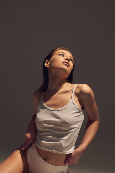 Πορτρέτο του νεαρού ελκυστική γυναίκα μοντέλο με καφέ μακριά σγουρά μαλλιά, σε λευκό φόρεμα bodysuit, μακριά πόδια με μαυρισμένο δέρμα, λεπτό σώμα, κάθεται στο παρασκήνιο με ήλιο σκληρό φως. Η έννοια μιας όμορφης κομψής μορφής, θετικού στο σώμα, υγιεινού τρόπου ζωής - Φωτογραφία, εικόνα