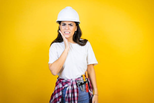 Young caucasian woman wearing hardhat and builder clothes over isolated yellow background touching mouth with hand with painful expression because of toothache or dental illness on teeth - Zdjęcie, obraz