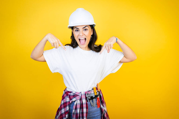Young caucasian woman wearing hardhat and builder clothes over isolated yellow background looking confident with smile on face, pointing oneself with fingers proud and happy. - Valokuva, kuva