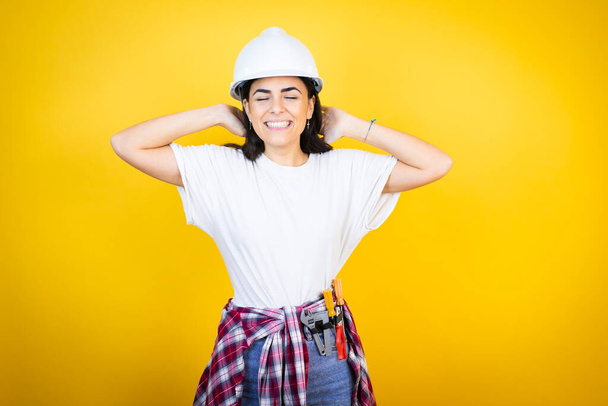 Young caucasian woman wearing hardhat and builder clothes over isolated yellow background relaxing and stretching, arms and hands behind head and neck smiling happy - Photo, Image